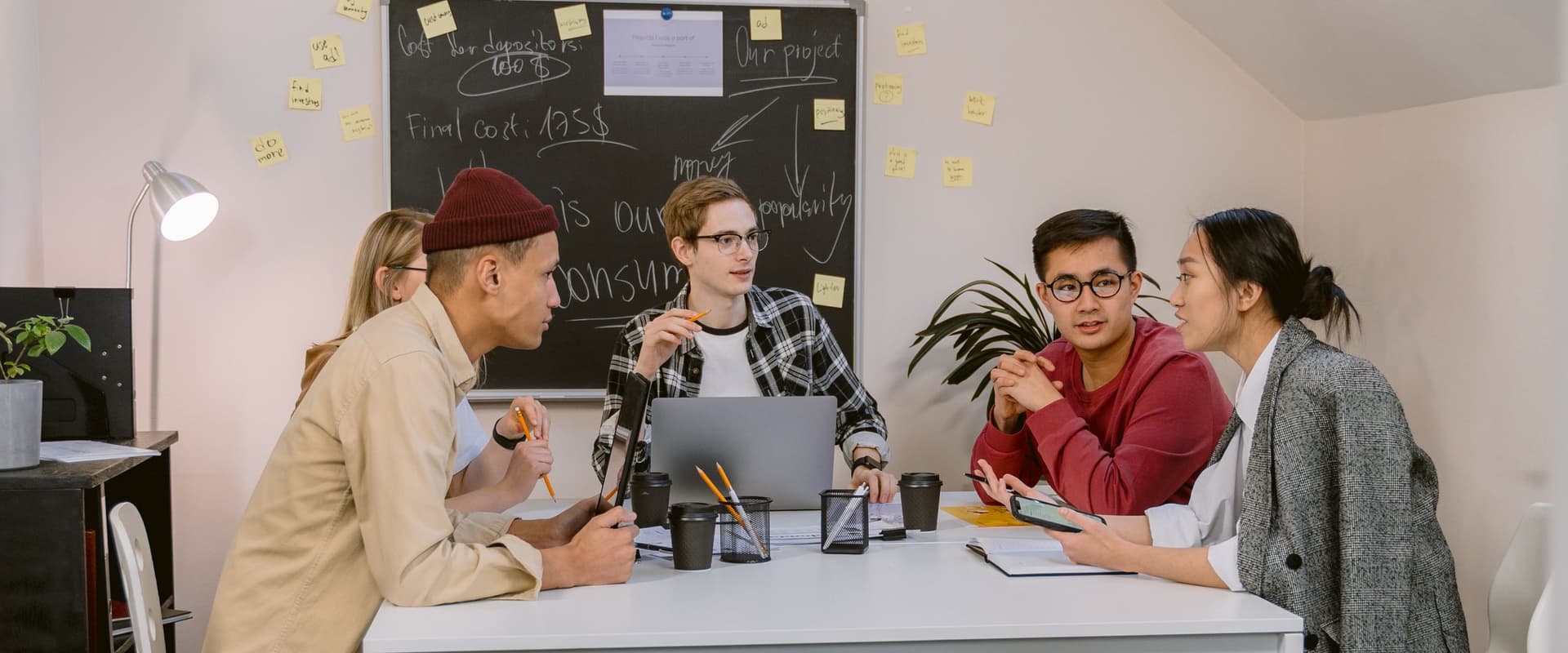 A small team talking around a white table in front of a blackboard covered in notes