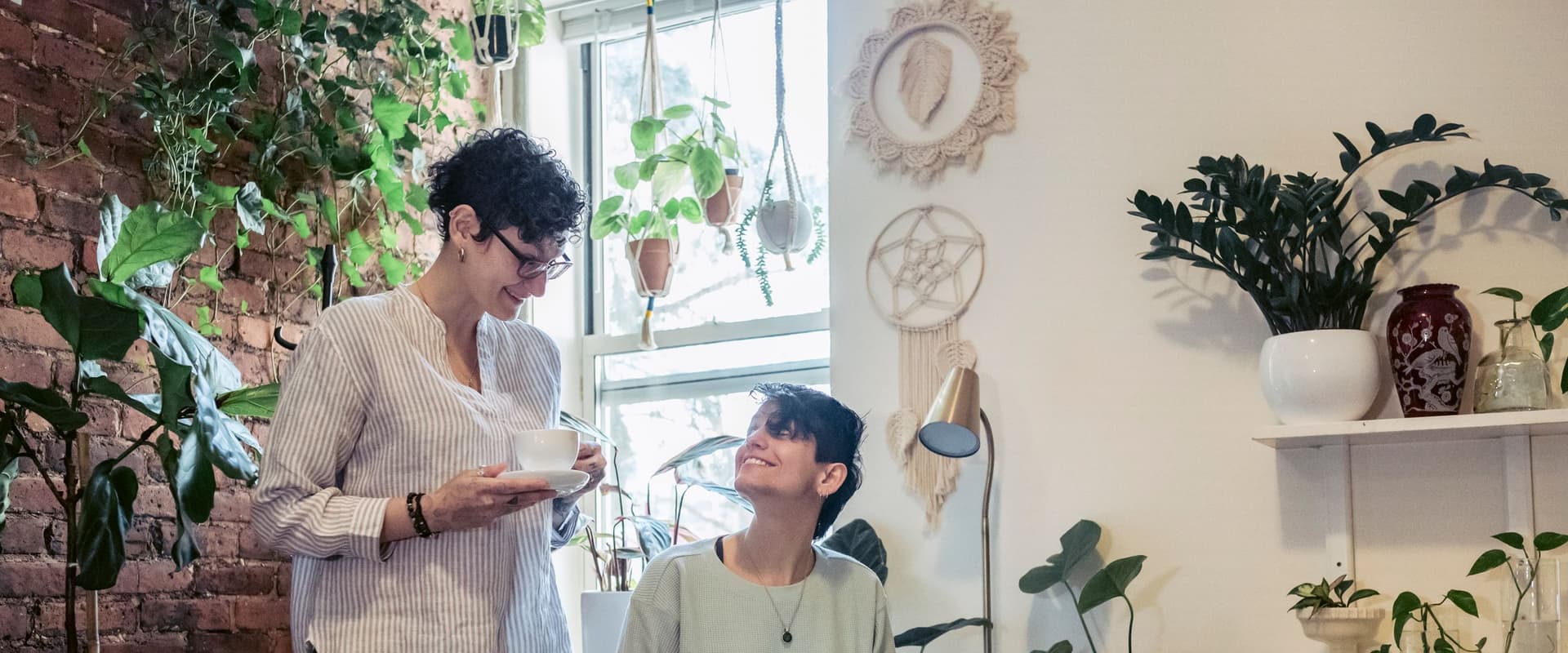 Two colleagues talking in a bright workspace full of plants, one standing with a cup and saucer and the other seated with a laptop