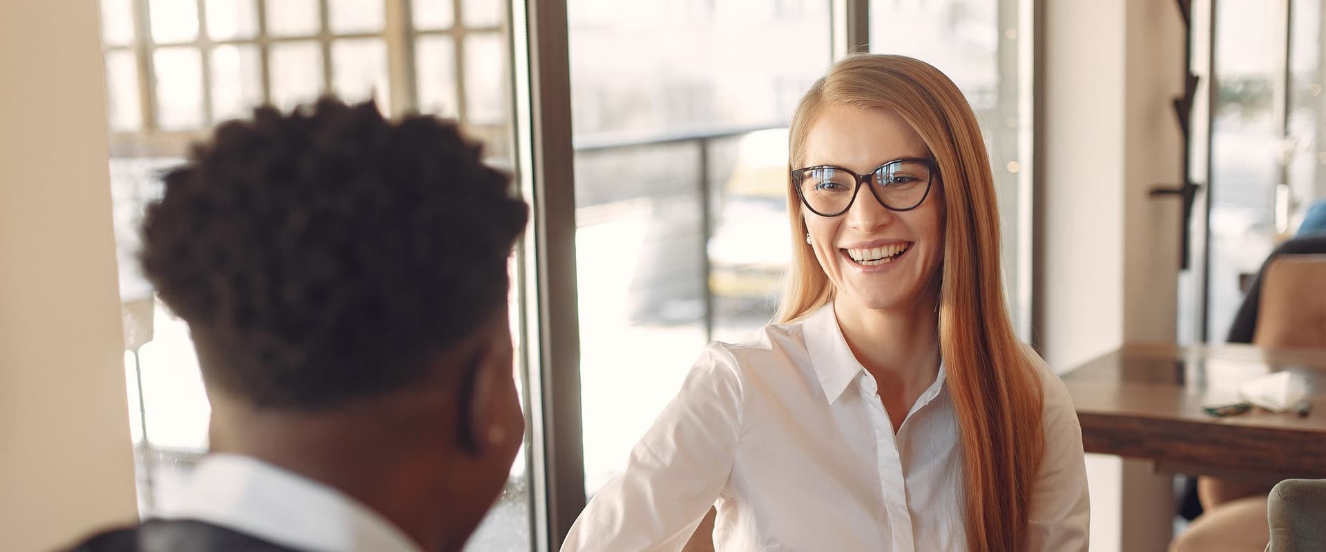 Two colleagues sharing a relaxed conversation over coffee at a table by a window, one smiling as she talks