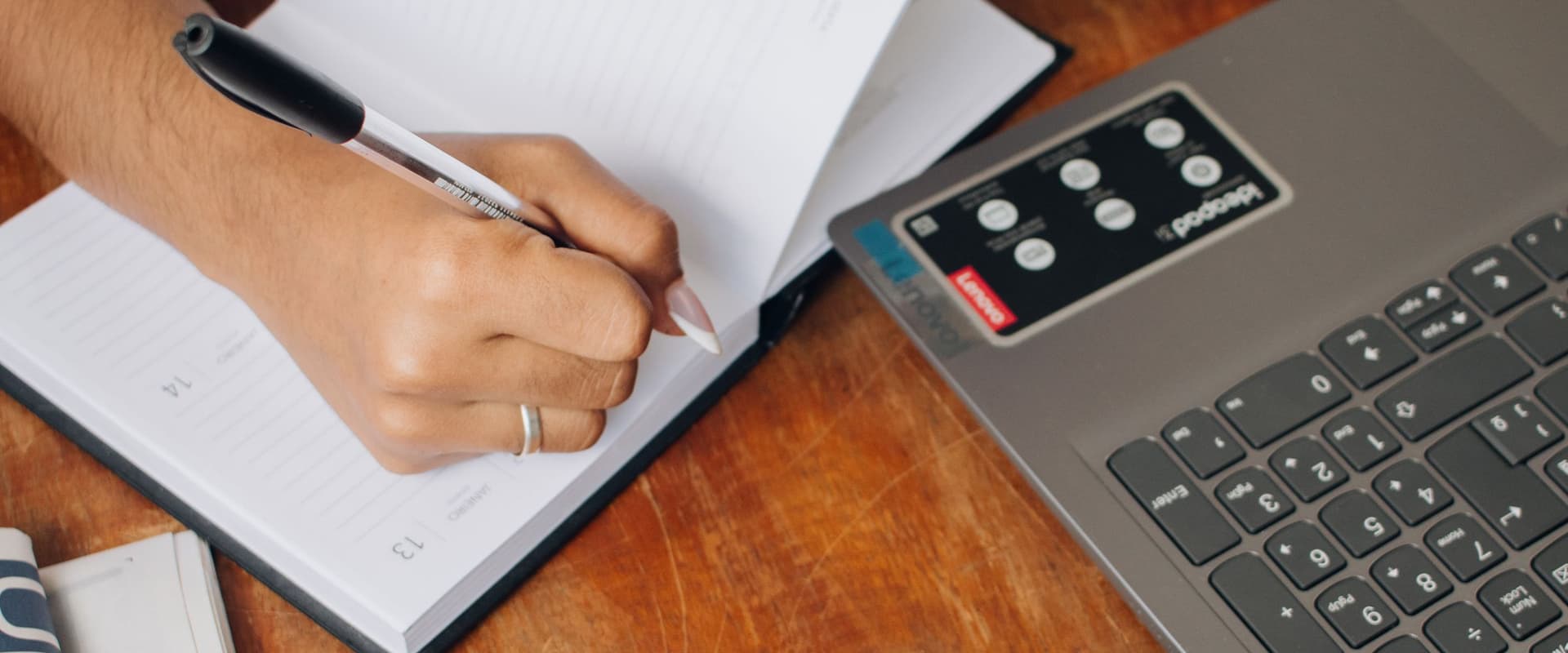 A hand writing notes in a notebook beside an open laptop