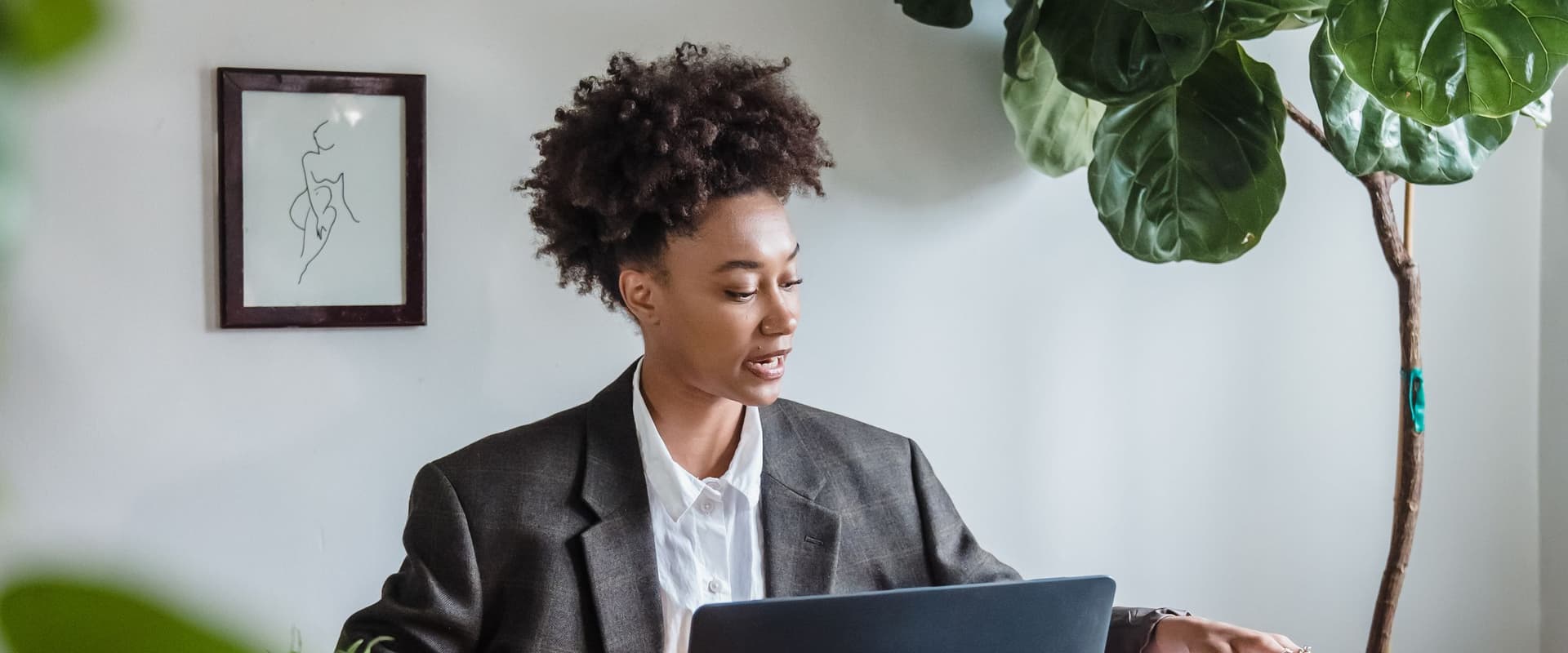 A woman in a grey blazer working at a laptop beside a large indoor plant